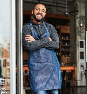a small business owner wearing a blue apron leaning against a glass door