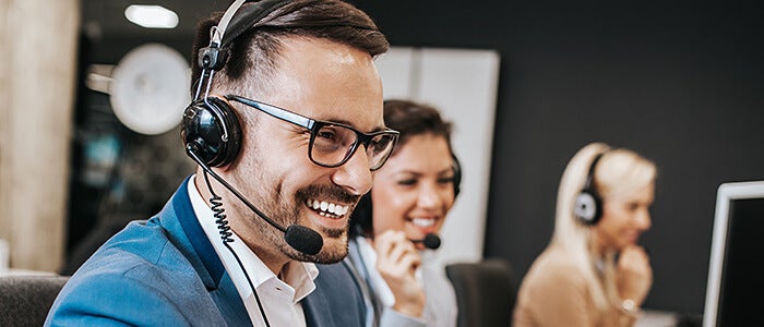 A man at the forefront of the image smiling and talking on a headset, with smiling women blurred behind him also with headsets.