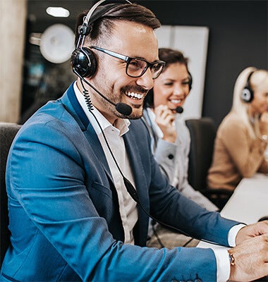 A man at the forefront of the image smiling and talking on a headset, with smiling women blurred behind him also with headsets.