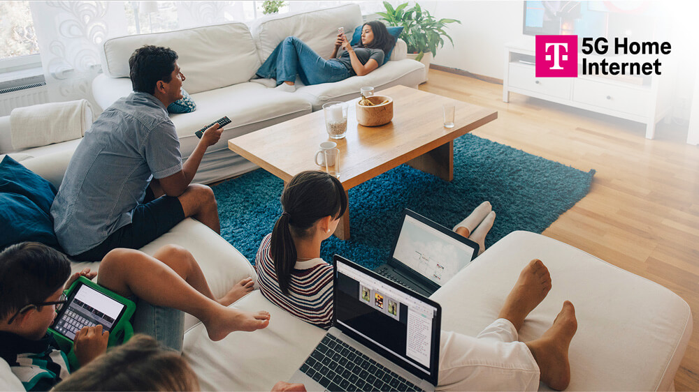 Family of five in living room using two laptops, a tablet, and a phone with T-Mobile 5G Home Internet.