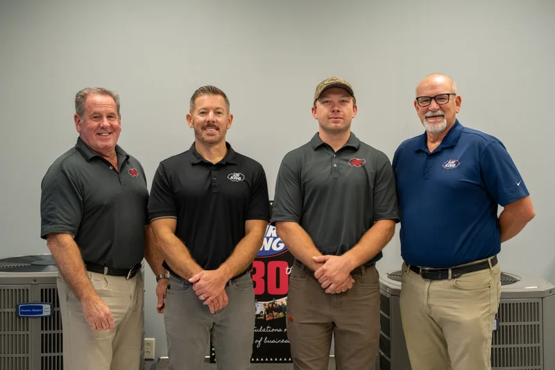 Four men in polo shirts stand side by side in front of air conditioning units.