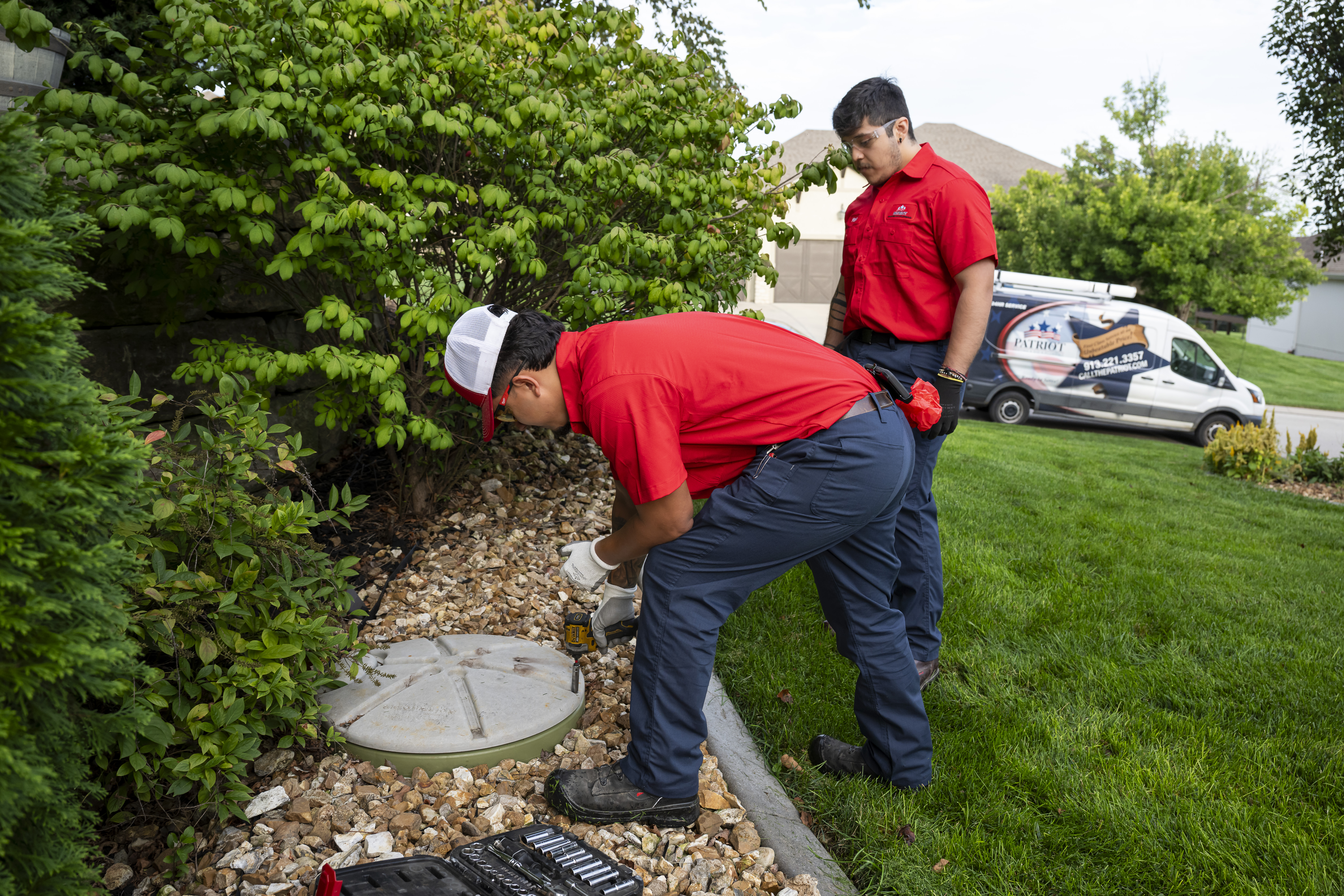 Patriot Heating & AC plumbers servicing an outdoor access lid with a Patriot service van in the background