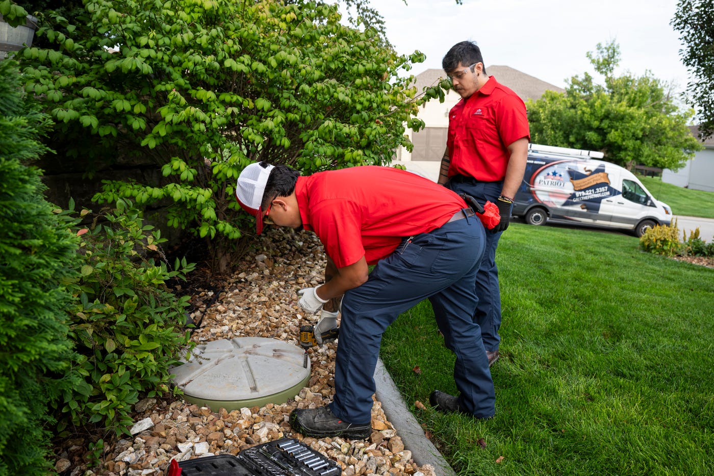 Patriot Heating & AC plumbers servicing an outdoor access lid with a Patriot service van in the background