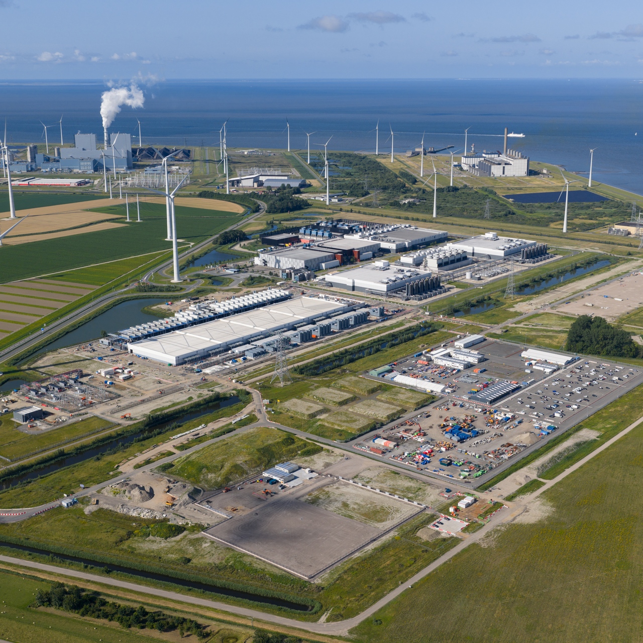 An aerial view of a large data center with wind turbines around it and other industrial buildings.