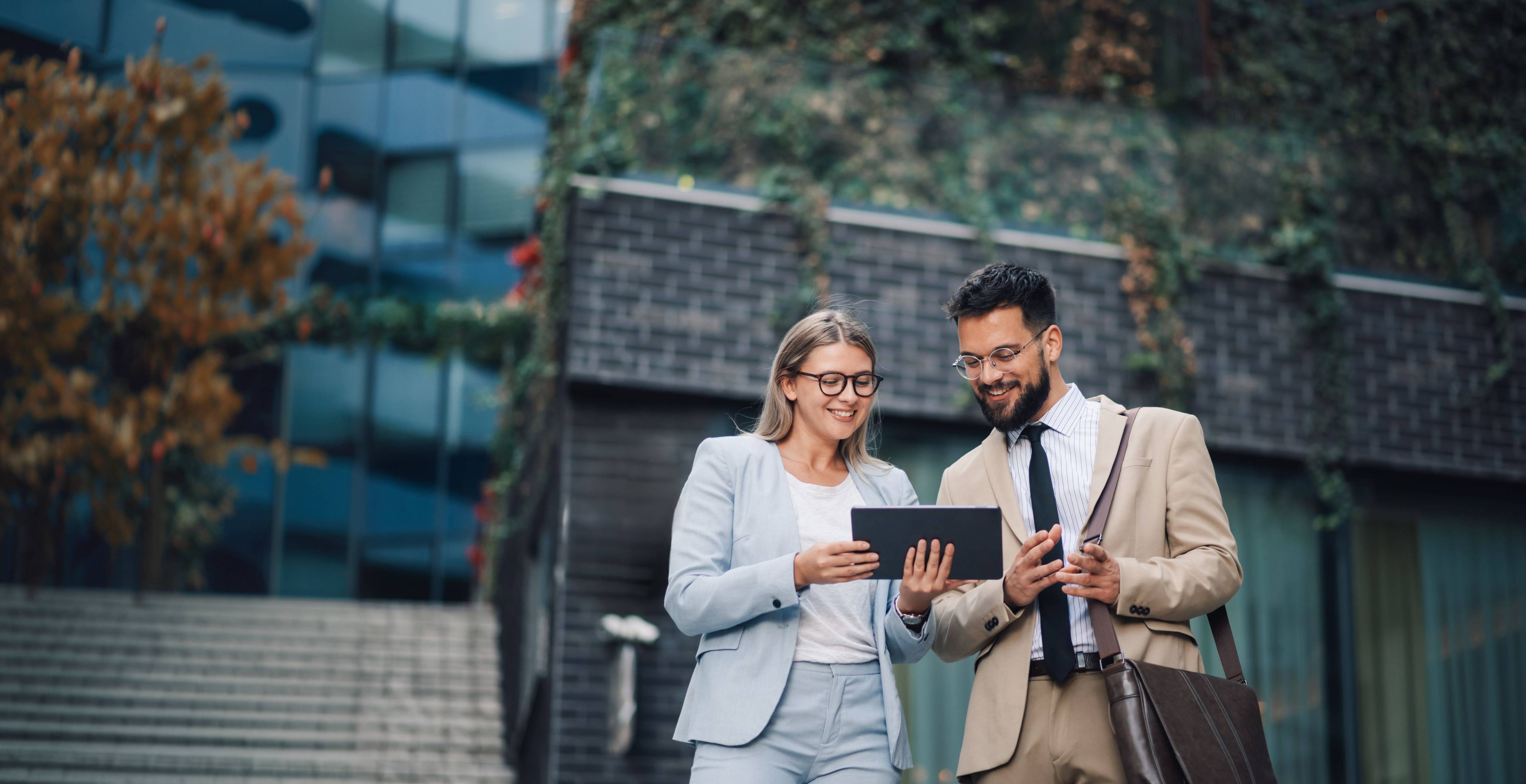 Two professional valuers are standing outside in front of an office building, smiling while looking at a digital tablet and discussing valuations