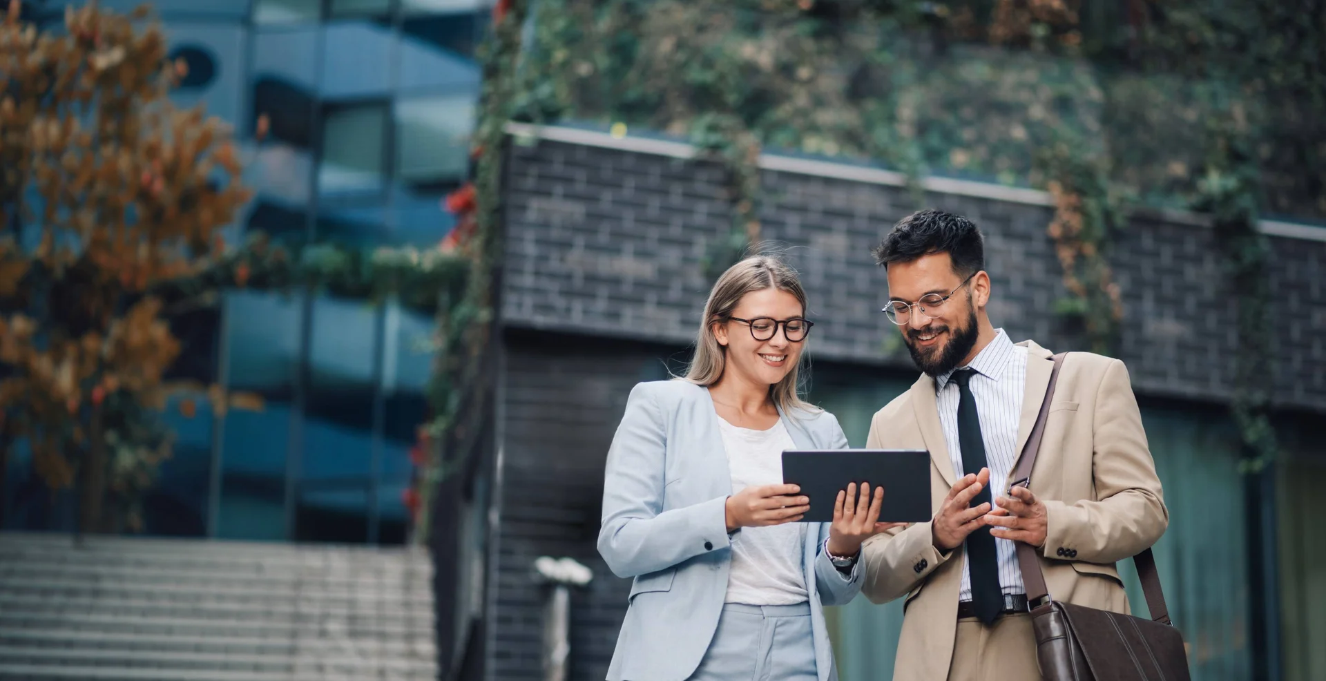 Two professional valuers are standing outside in front of an office building, smiling while looking at a digital tablet and discussing valuations