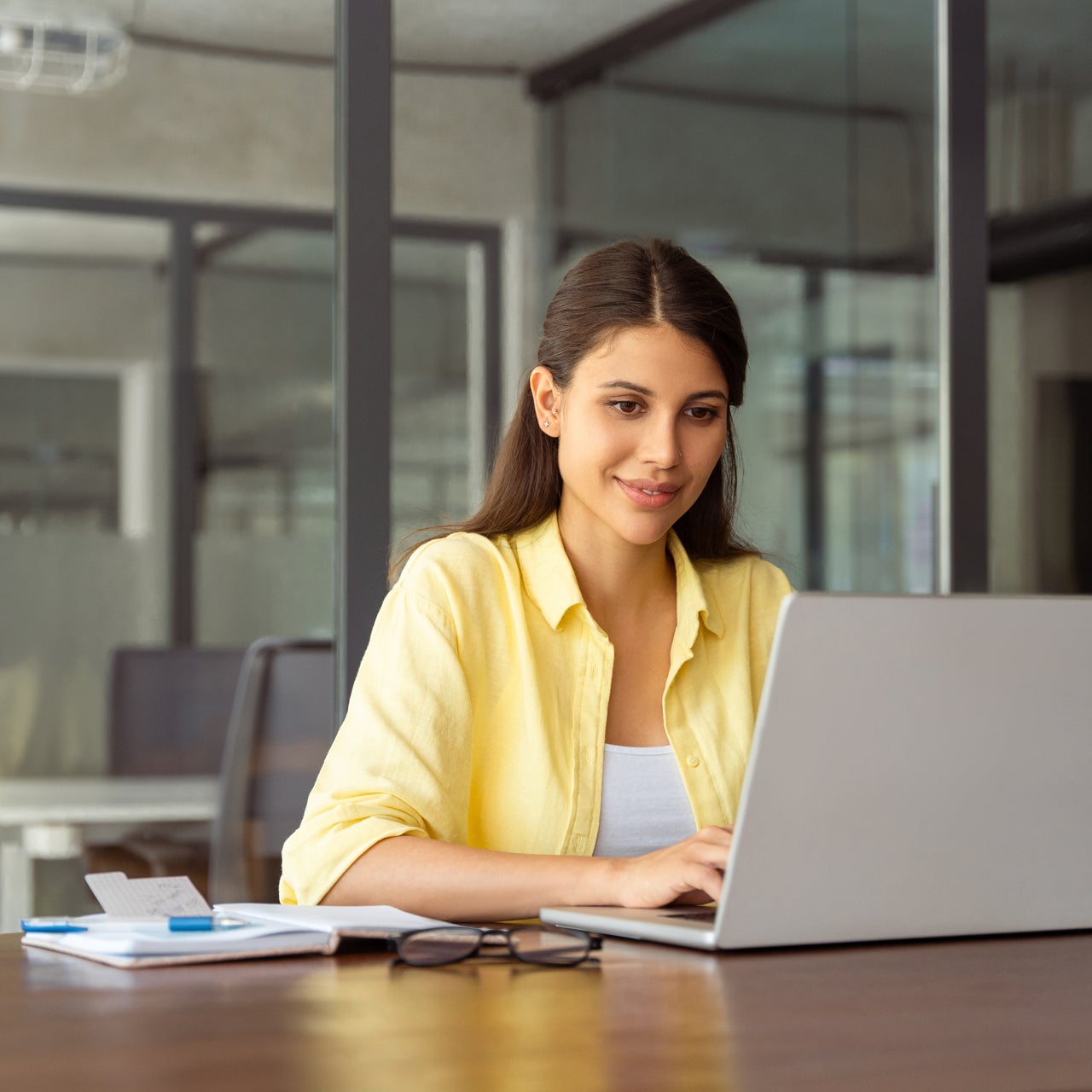 A young woman sits at a desk and smiles while working on a silver laptop.