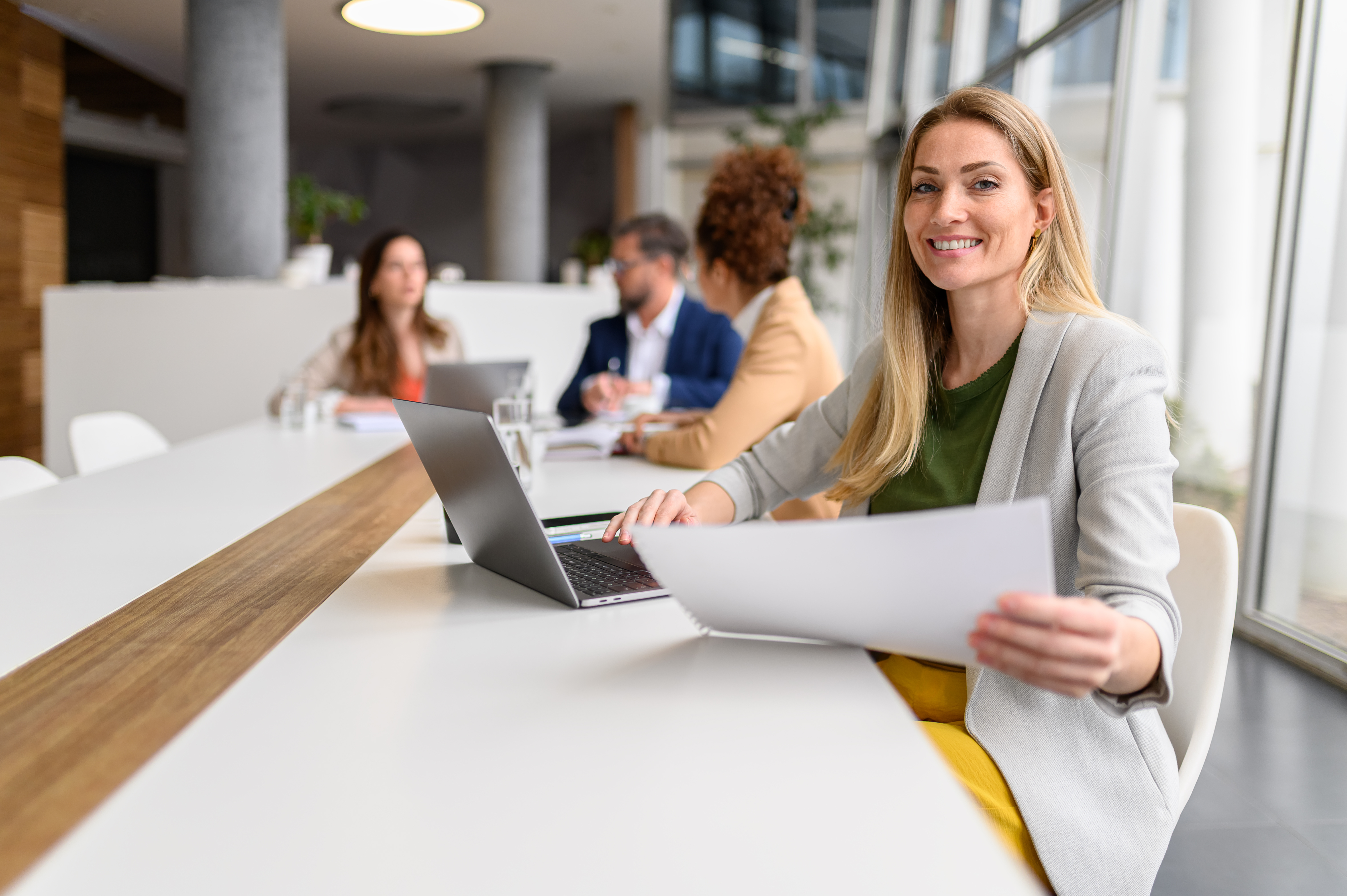 A smiling businesswoman in a grey blazer is seen holding papers.