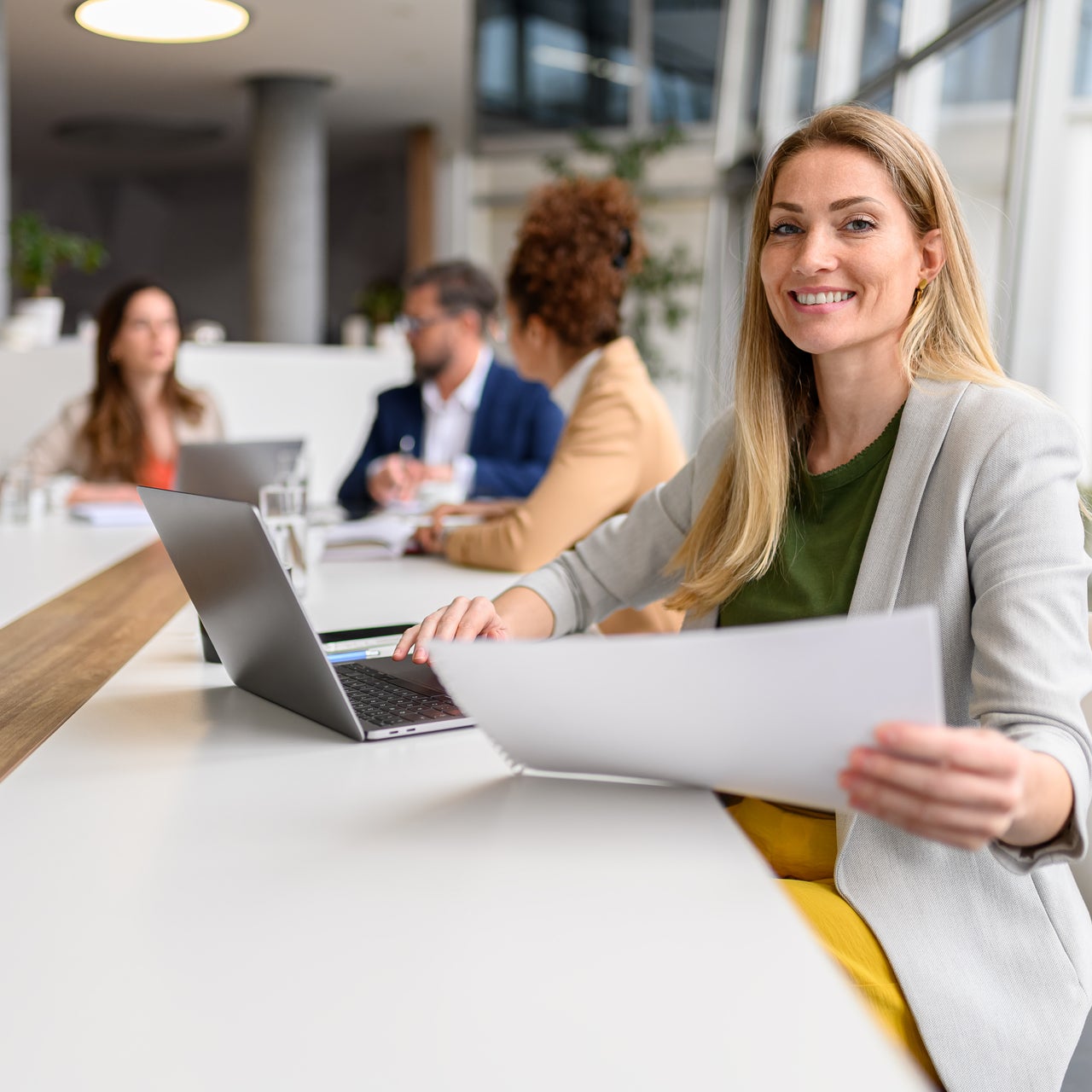 A smiling businesswoman in a grey blazer is seen holding papers.