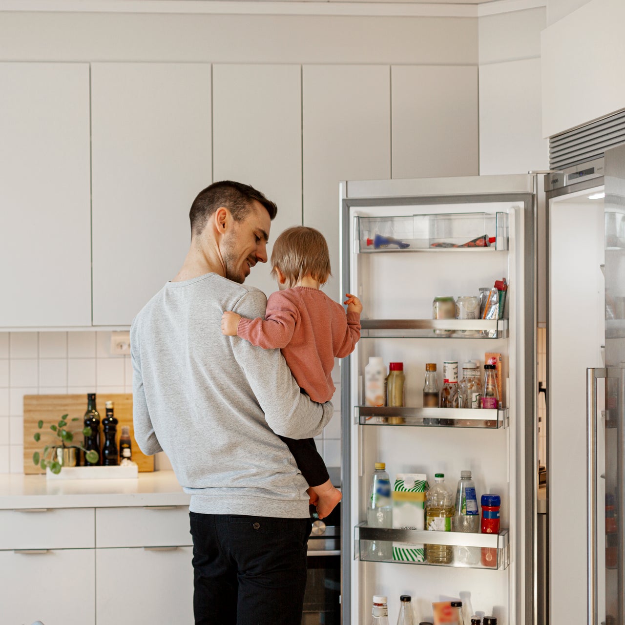 A young father holding his infant in his lap is standing in front of an open refrigerator.