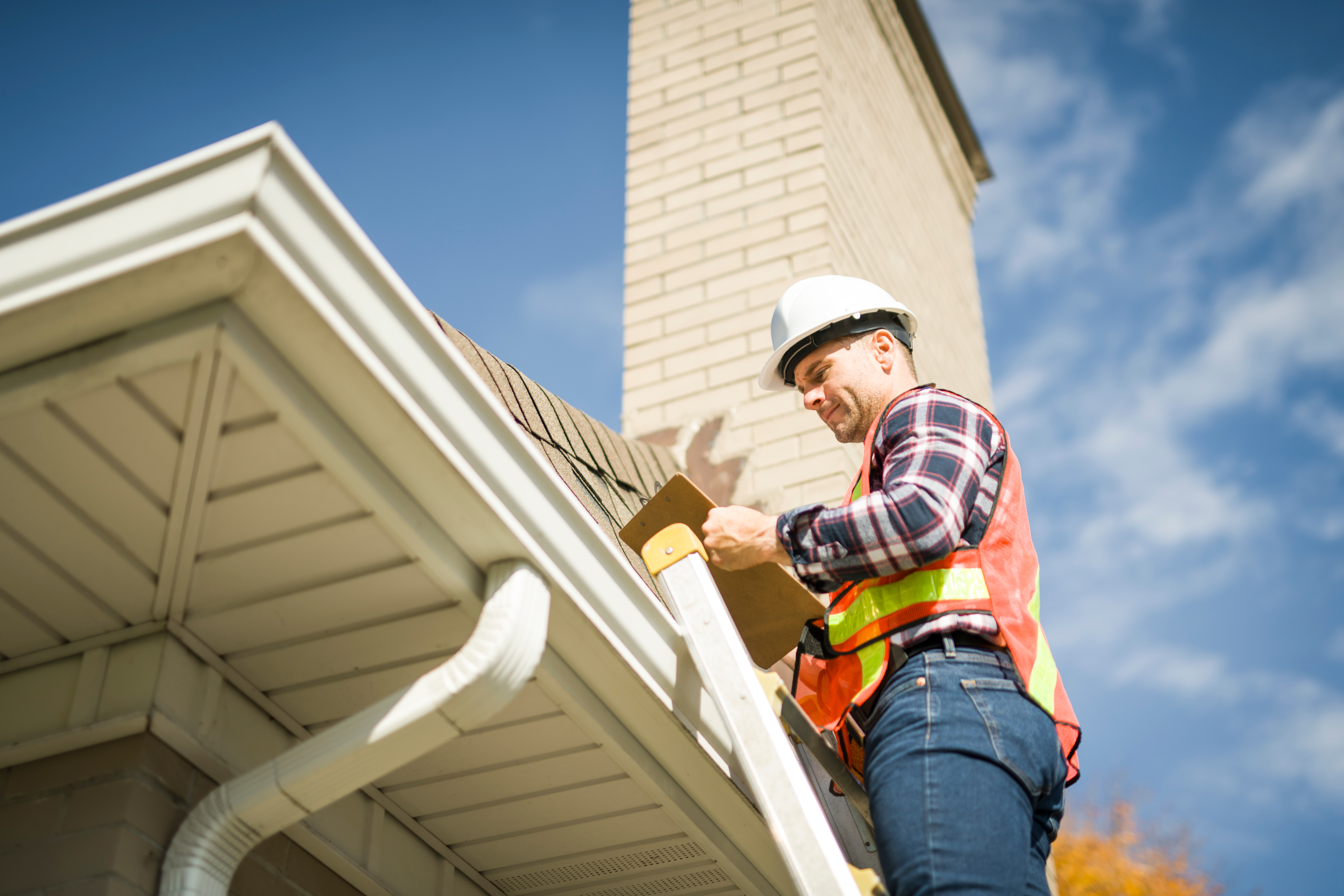 A man wearing property safter is standing on a ladder, inspecting a roof and gutter system.