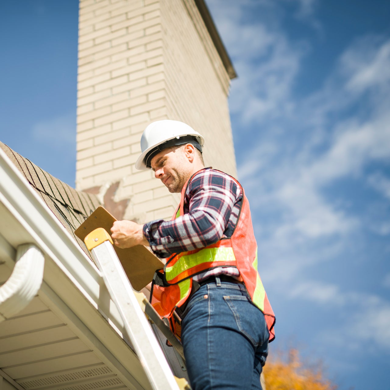 A man wearing property safter is standing on a ladder, inspecting a roof and gutter system.