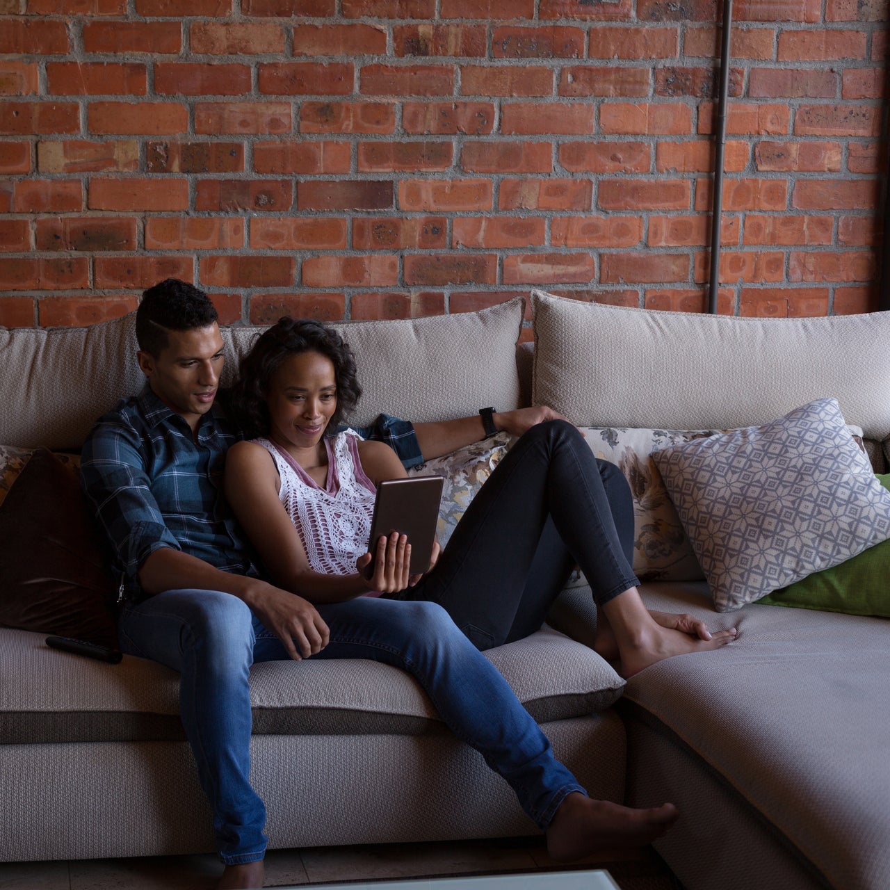 A man and a woman are sitting together on a couch, looking at a tablet. The man is wearing a blue plaid shirt and jeans, while the woman is wearing a white tank top and black leggings. They are both barefoot. The couch is beige and there is a brick wall behind them. There are also several pillows on the couch. The lighting is soft and natural, creating a relaxed and intimate atmosphere.