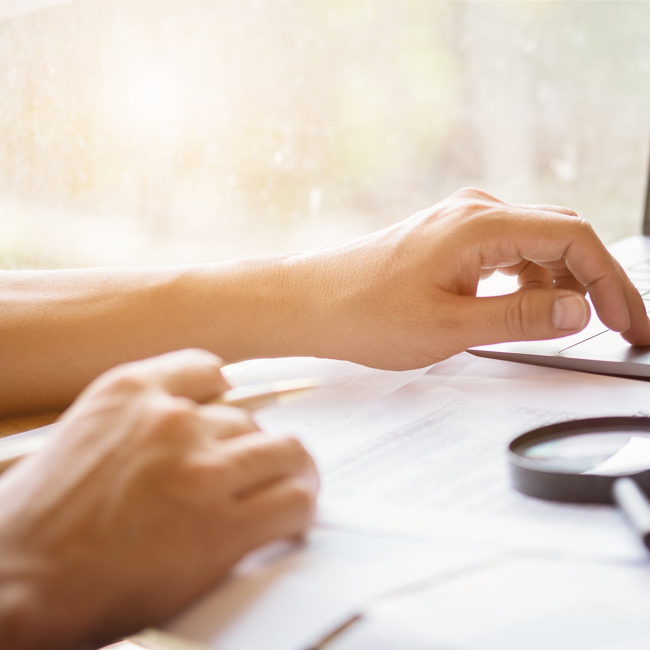 A hand is visible on a desk full of papers and a magnifying glass.