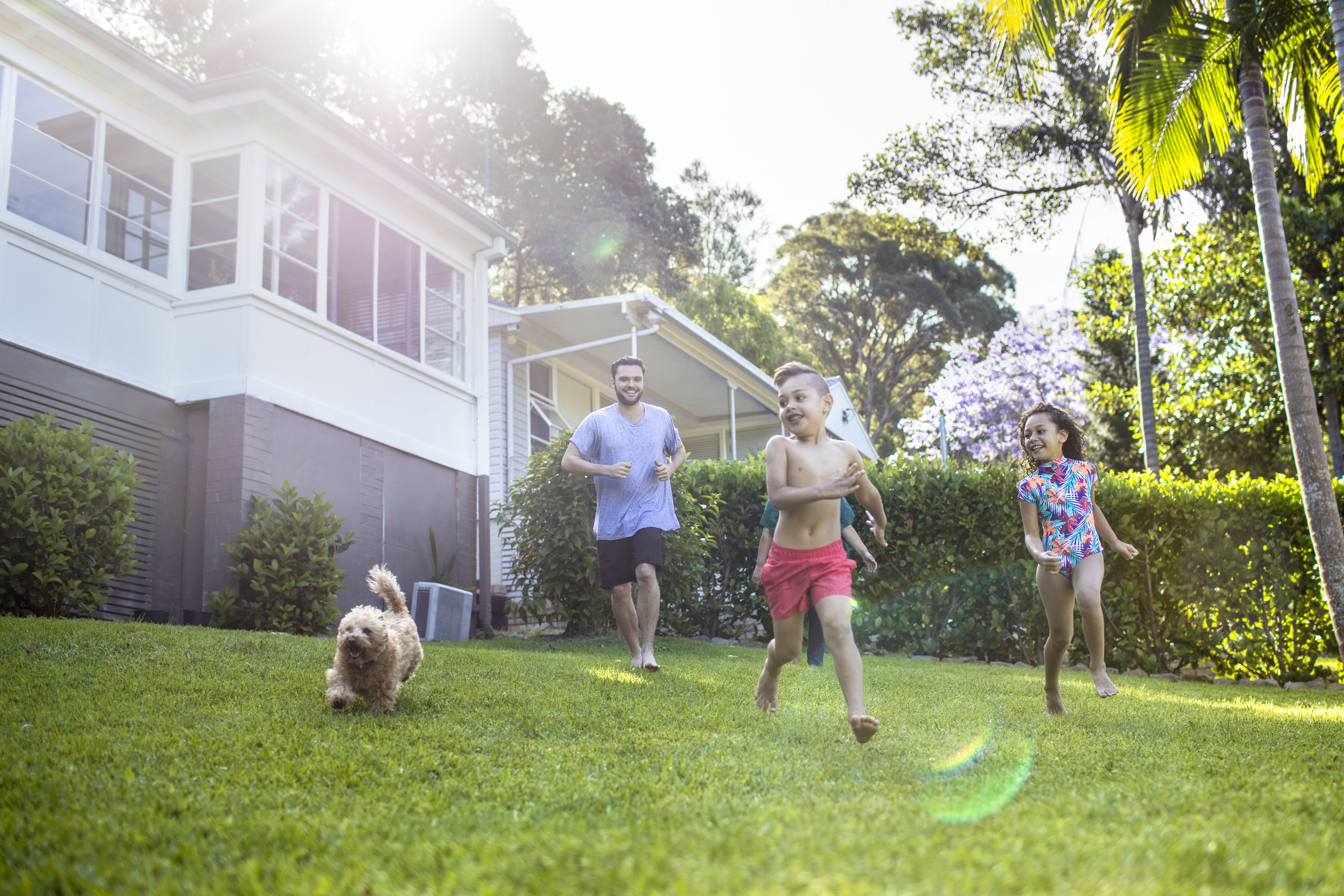 A man and three children are running barefoot on a green lawn in a backyard.