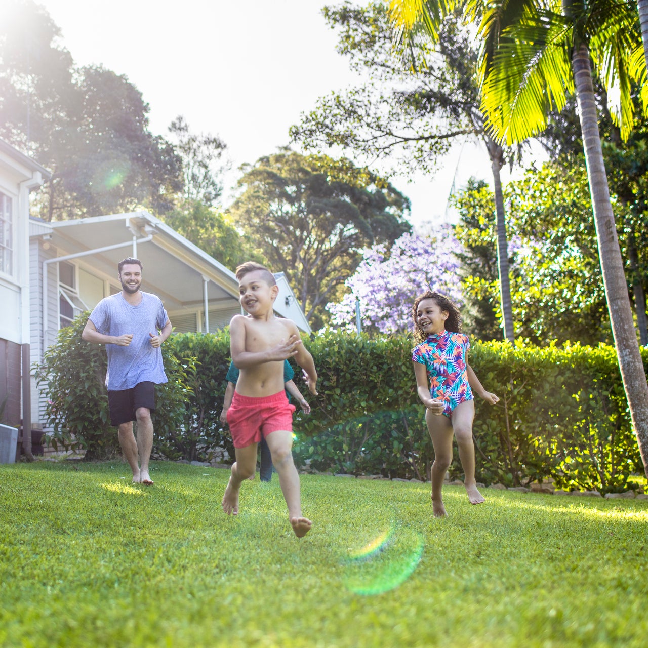 A man and three children are running barefoot on a green lawn in a backyard.
