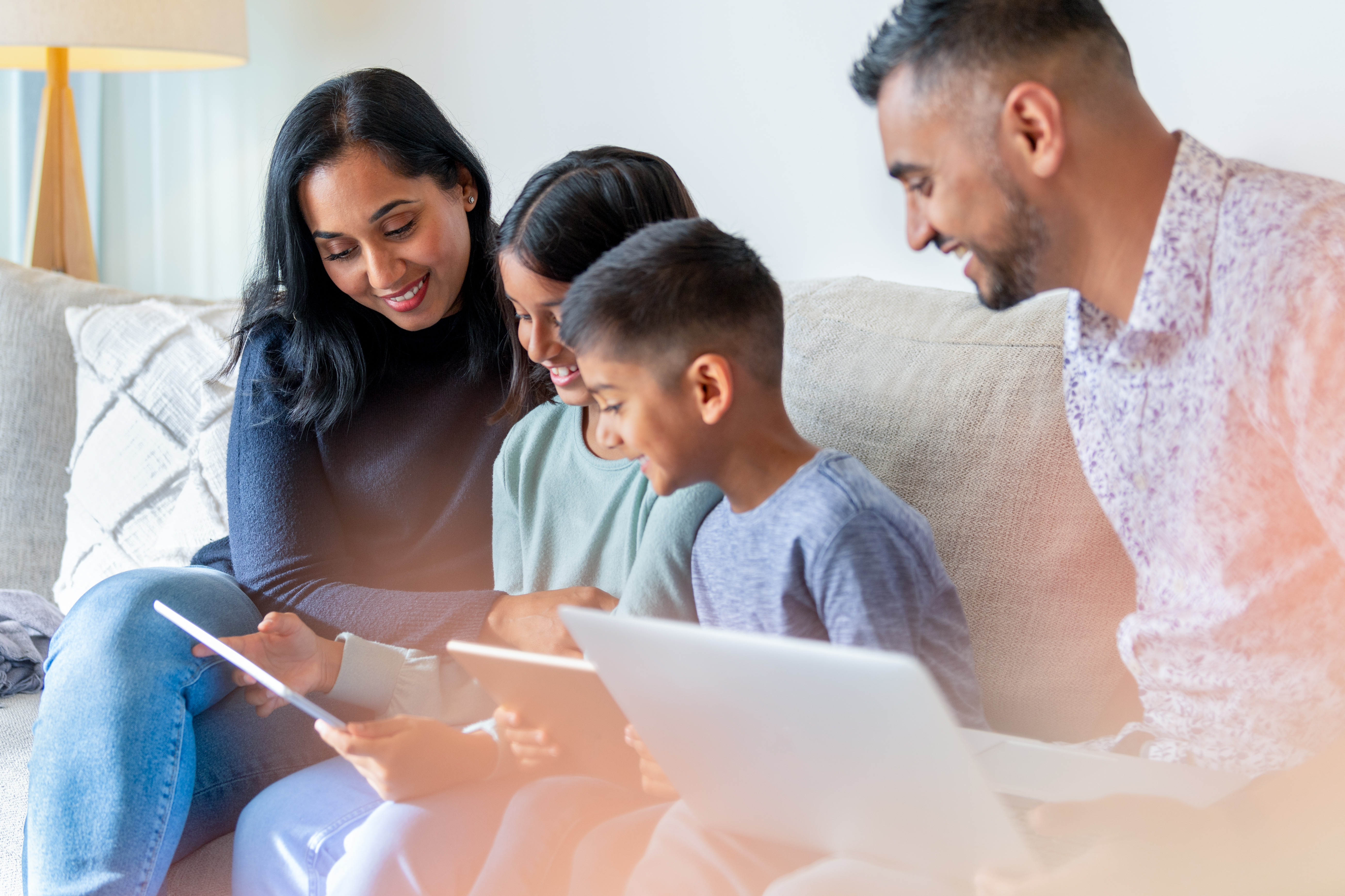 Two parents and two children on a couch in their house smiling while looking at a laptop and tablet.