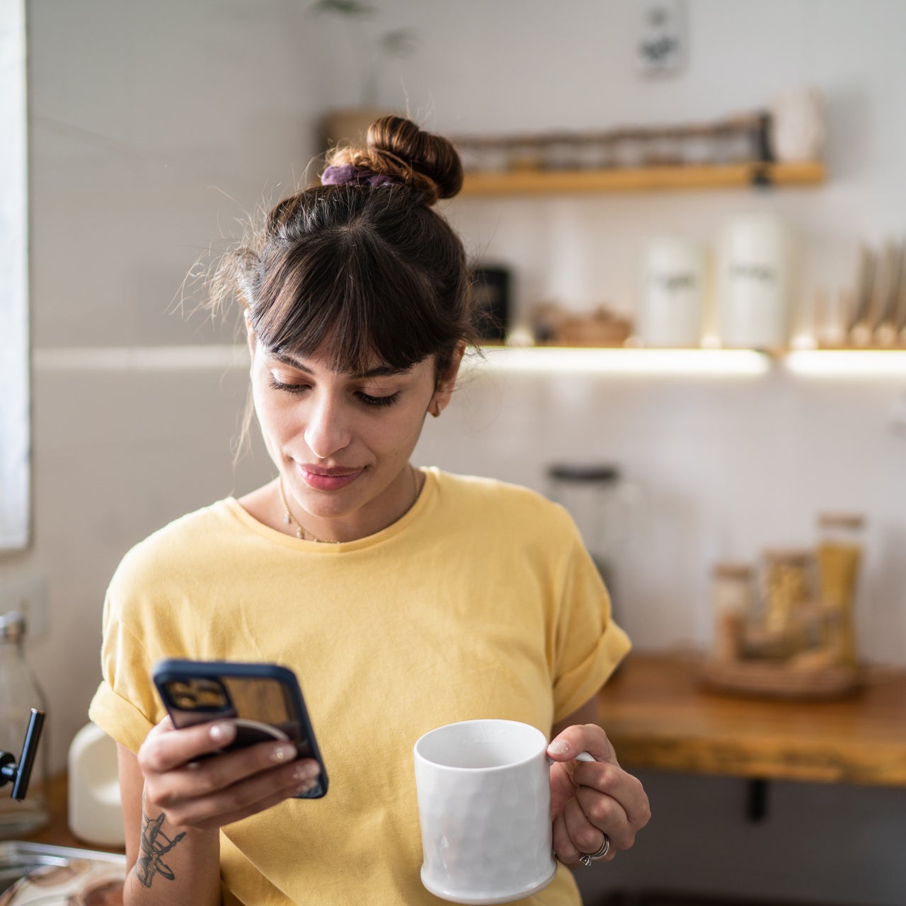 A young woman is seen holding a white mug in her right hand and a smartphone in her left hand.