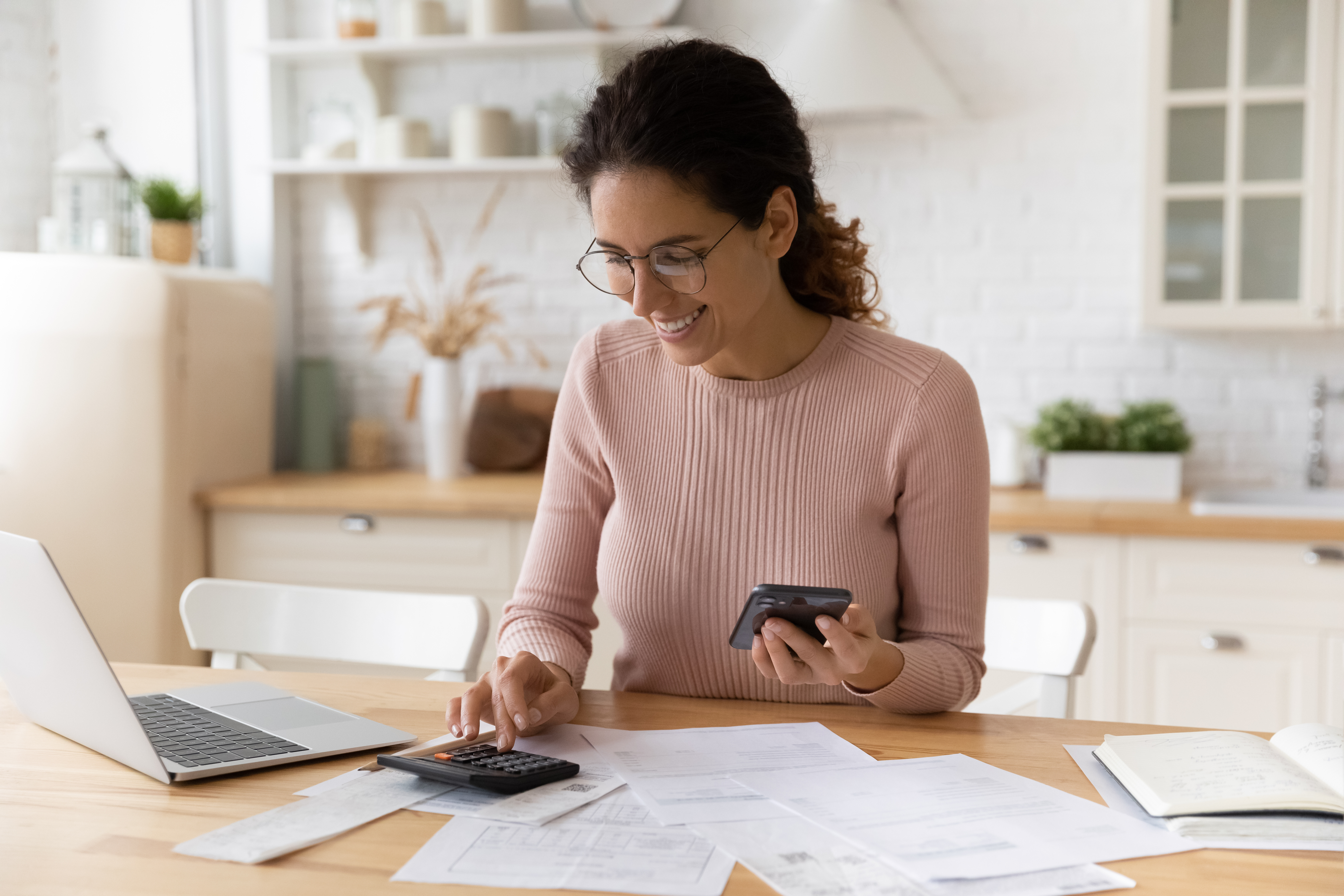 A woman smilingly working on her desk with calculator on hand and a mobile phone on the other. 