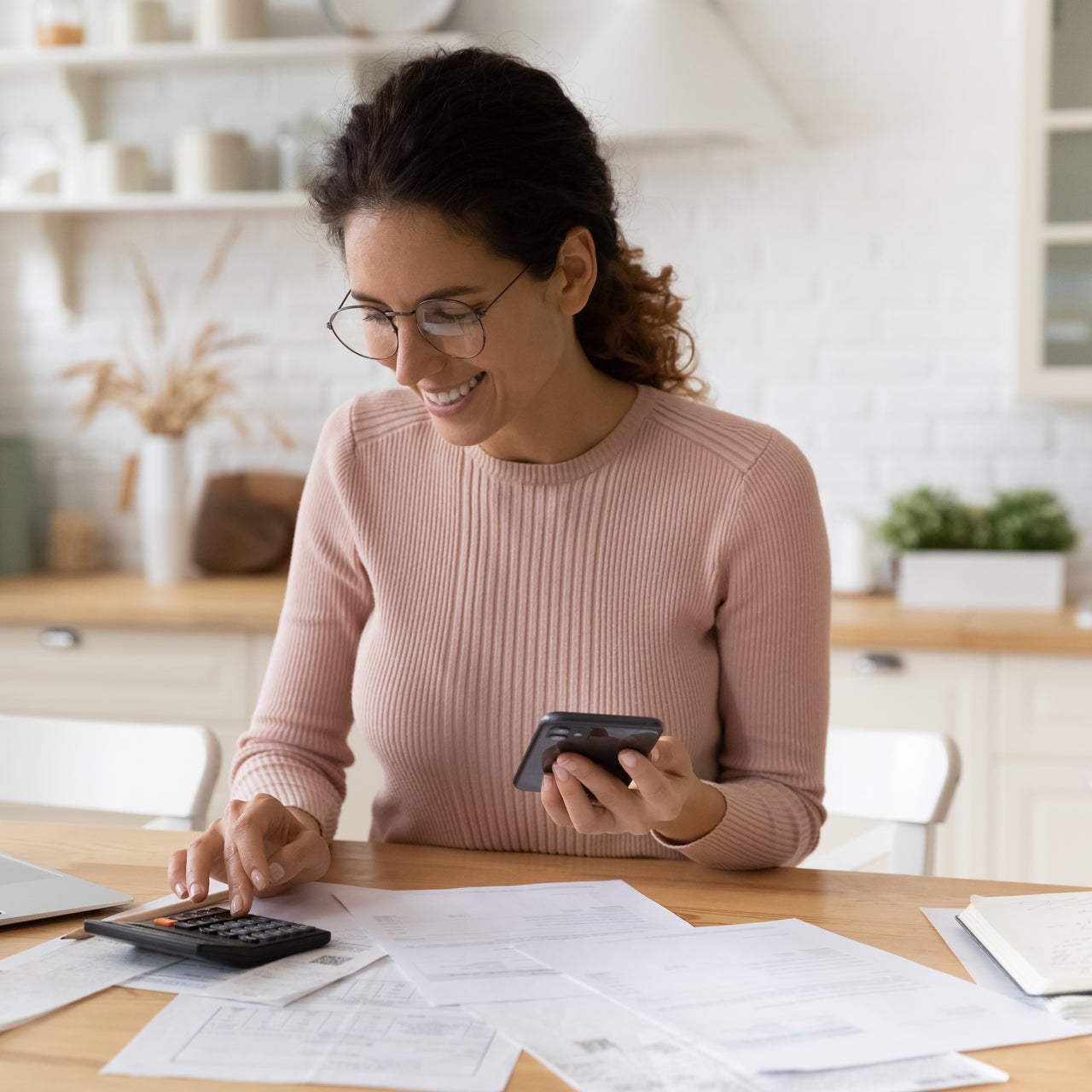 A woman smilingly working on her desk with calculator on hand and a mobile phone on the other.