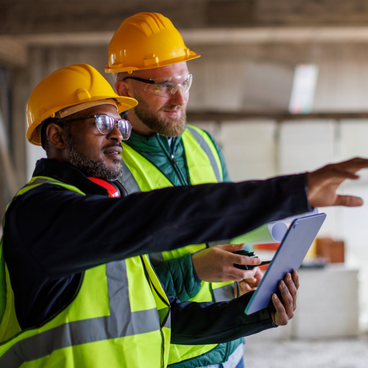 Two construction workers are wearing yellow hard hats and safety vests, one holding a tablet and the other with a walkie-talkie, seemingly discussing plans or progress on a construction site.