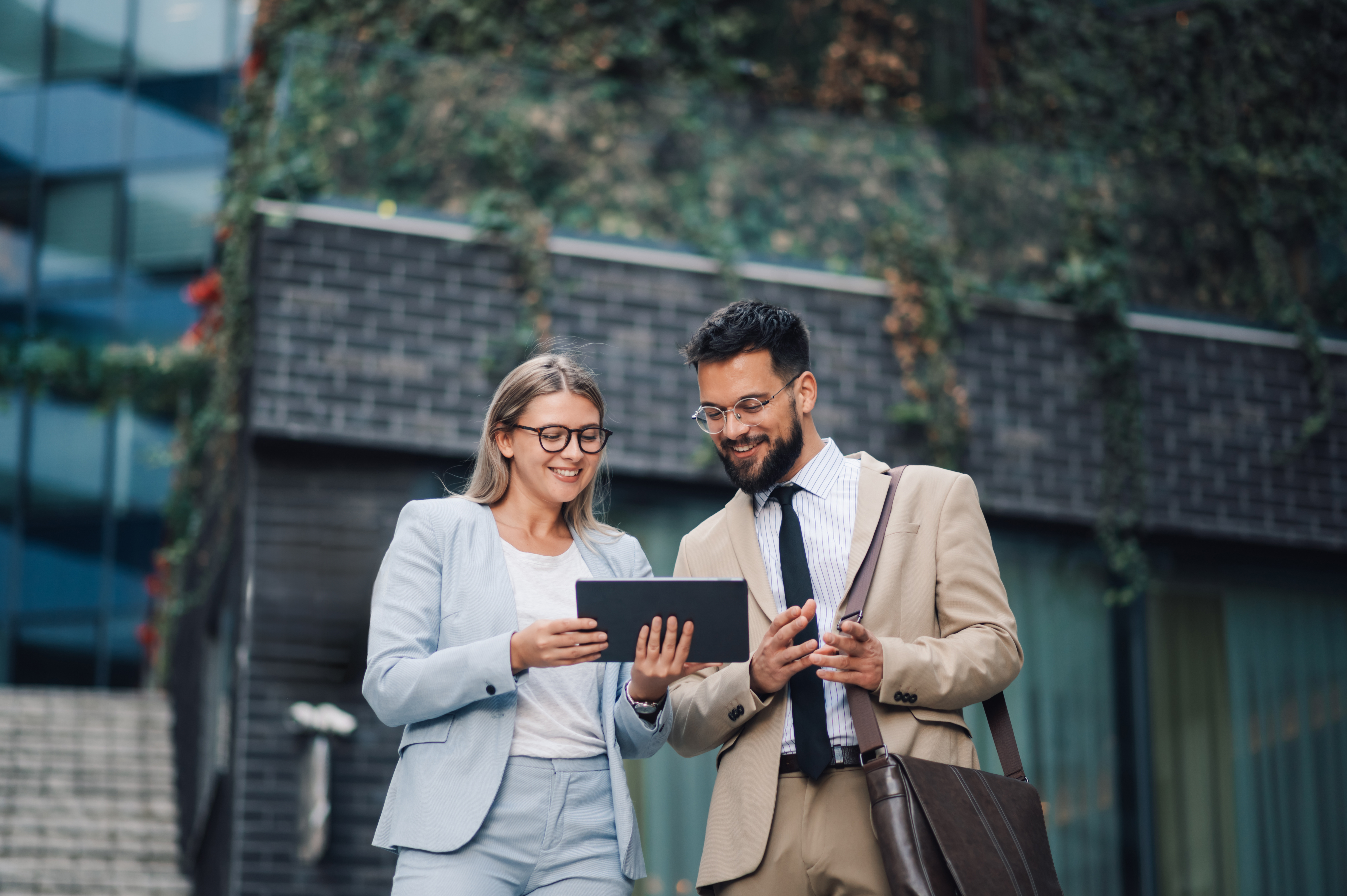 Two business people are standing outside in front of an office building, smiling while looking at a digital tablet