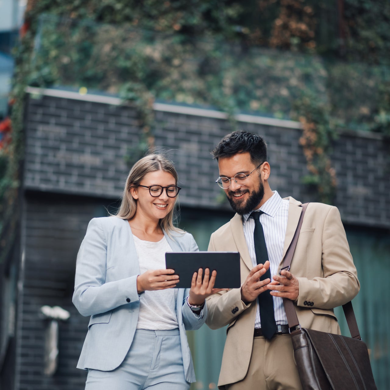 Two business people are standing outside in front of an office building, smiling while looking at a digital tablet