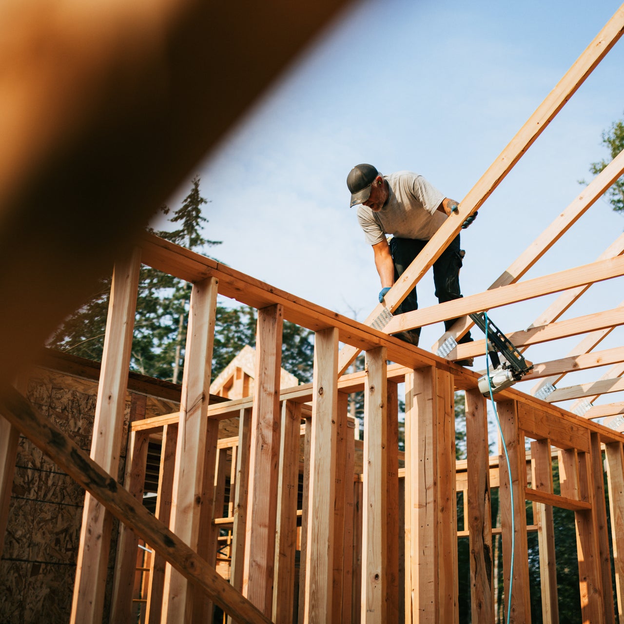 A construction worker is framing a house. The house frame is made of light-colored wood and is set against a backdrop of trees and a blue sky.