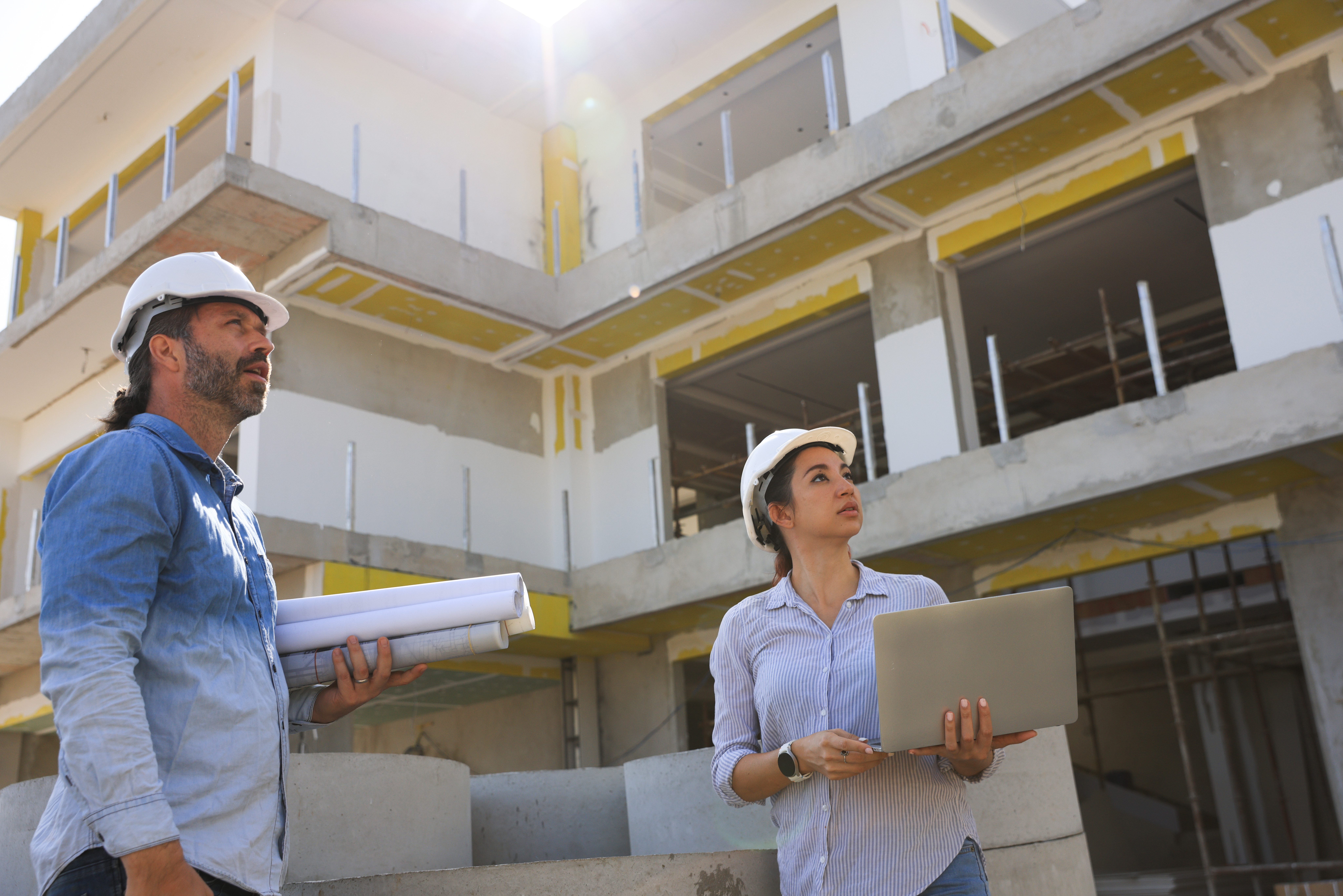 A woman is seen holding a laptop and blueprints at a construction site. The background shows an unfinished building. 