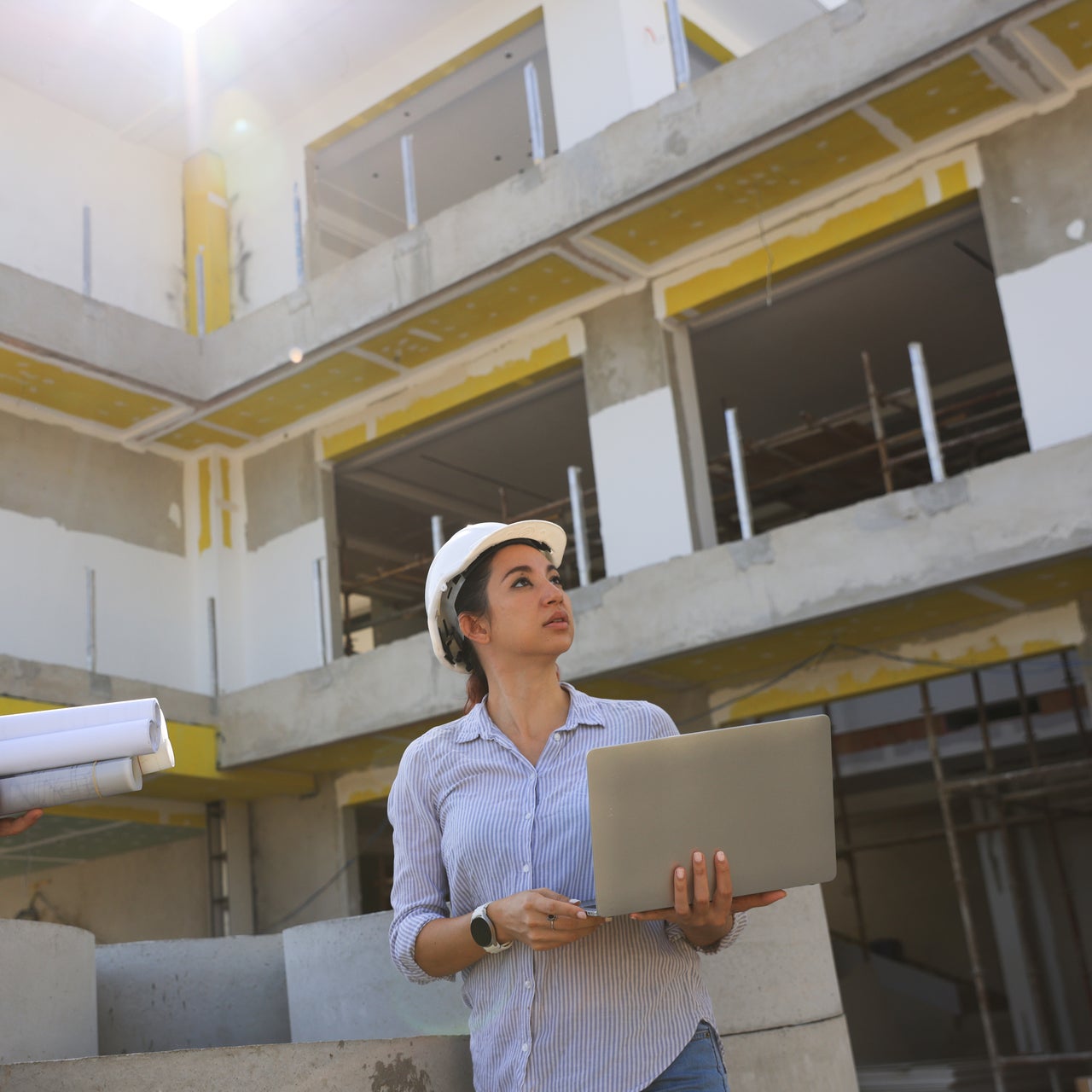 A woman is seen holding a laptop and blueprints at a construction site. The background shows an unfinished building.
