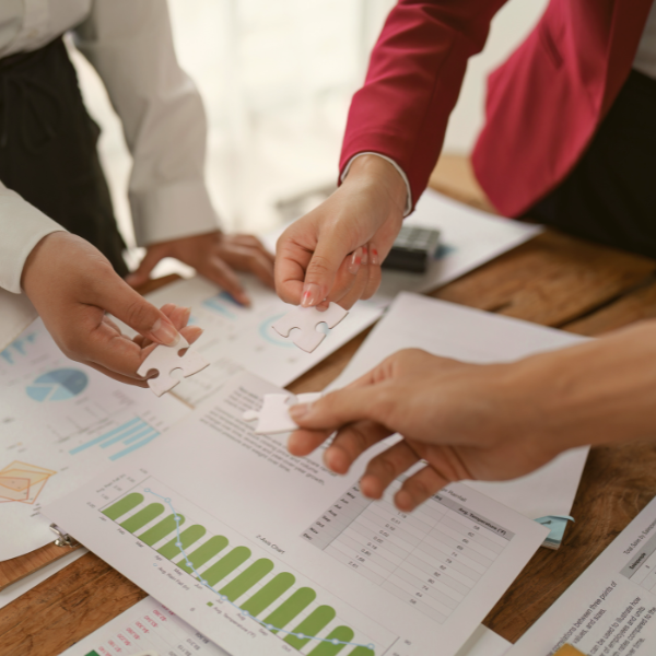 Three people piecing together jigsaw puzzle pieces over documents and charts.