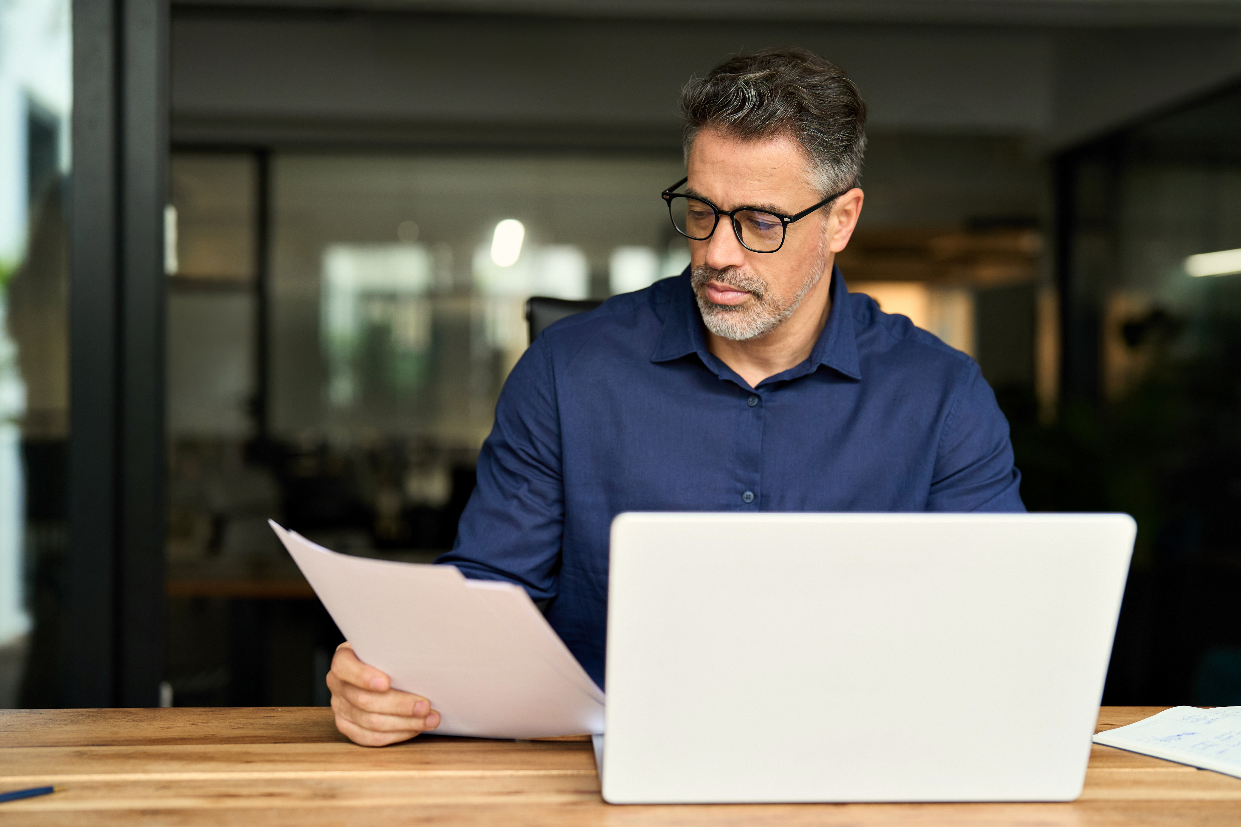 A middle aged person in formal attire is sitting on a desk with laptop screen open. 