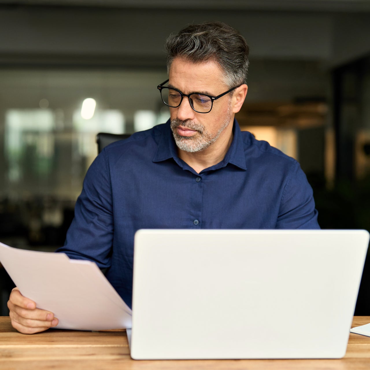 A middle aged person in formal attire is sitting on a desk with laptop screen open.