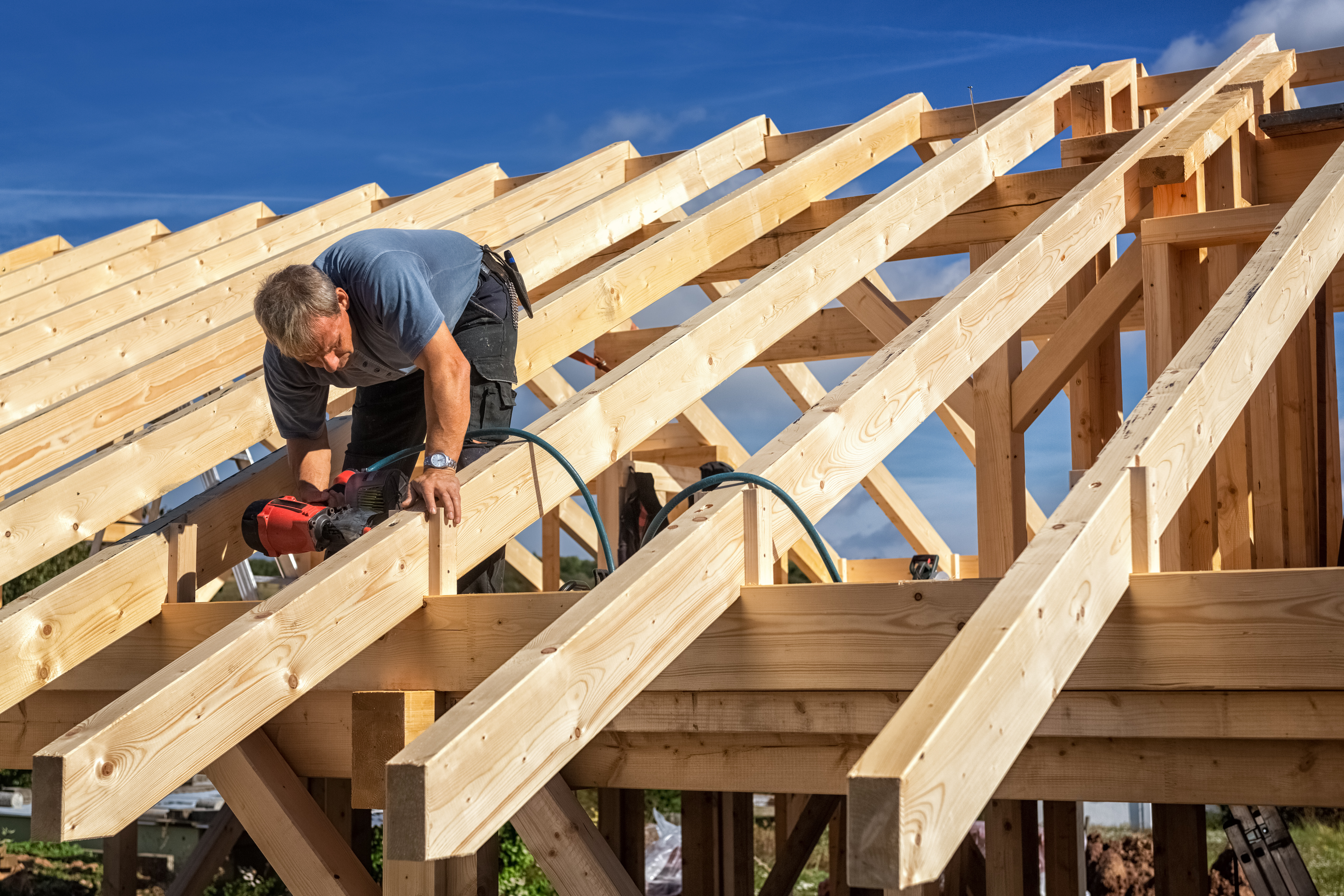 A construction worker on a roof frame, using a nail gun to secure wooden beams.
