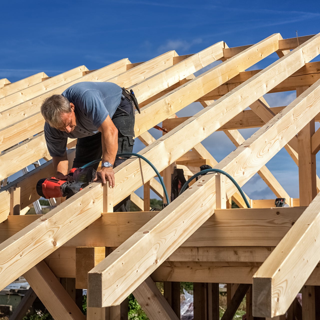 A construction worker on a roof frame, using a nail gun to secure wooden beams.