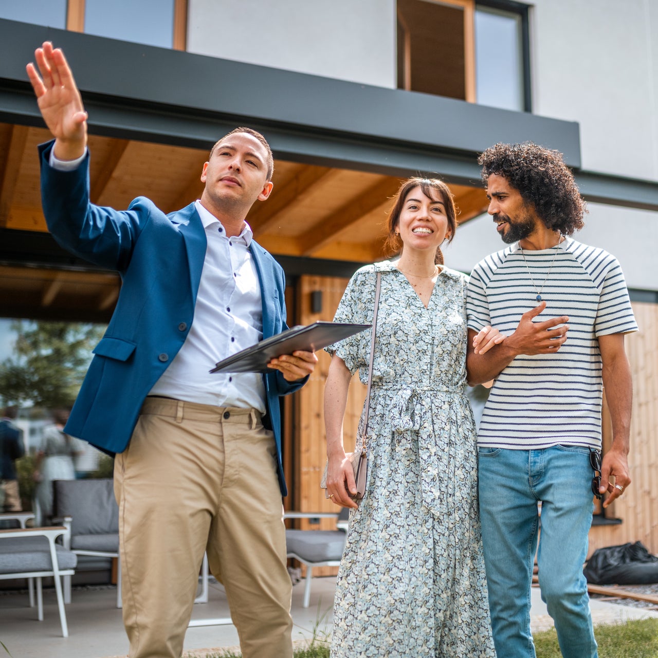 Three people stand outside a modern house, with one person holding a tablet and gesturing, discussing property details.