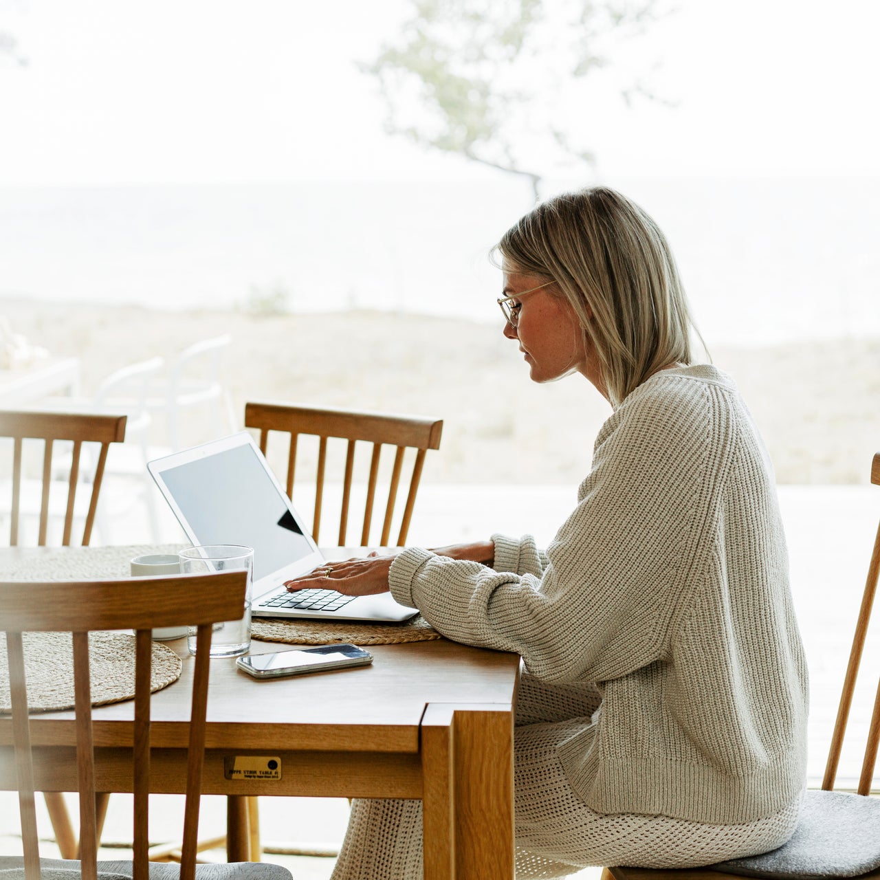 A woman sitting at a wooden table, working on a laptop. The table also has a smartphone and a glass of water on it. The woman has short blonde hair and is wearing glasses and a beige sweater.