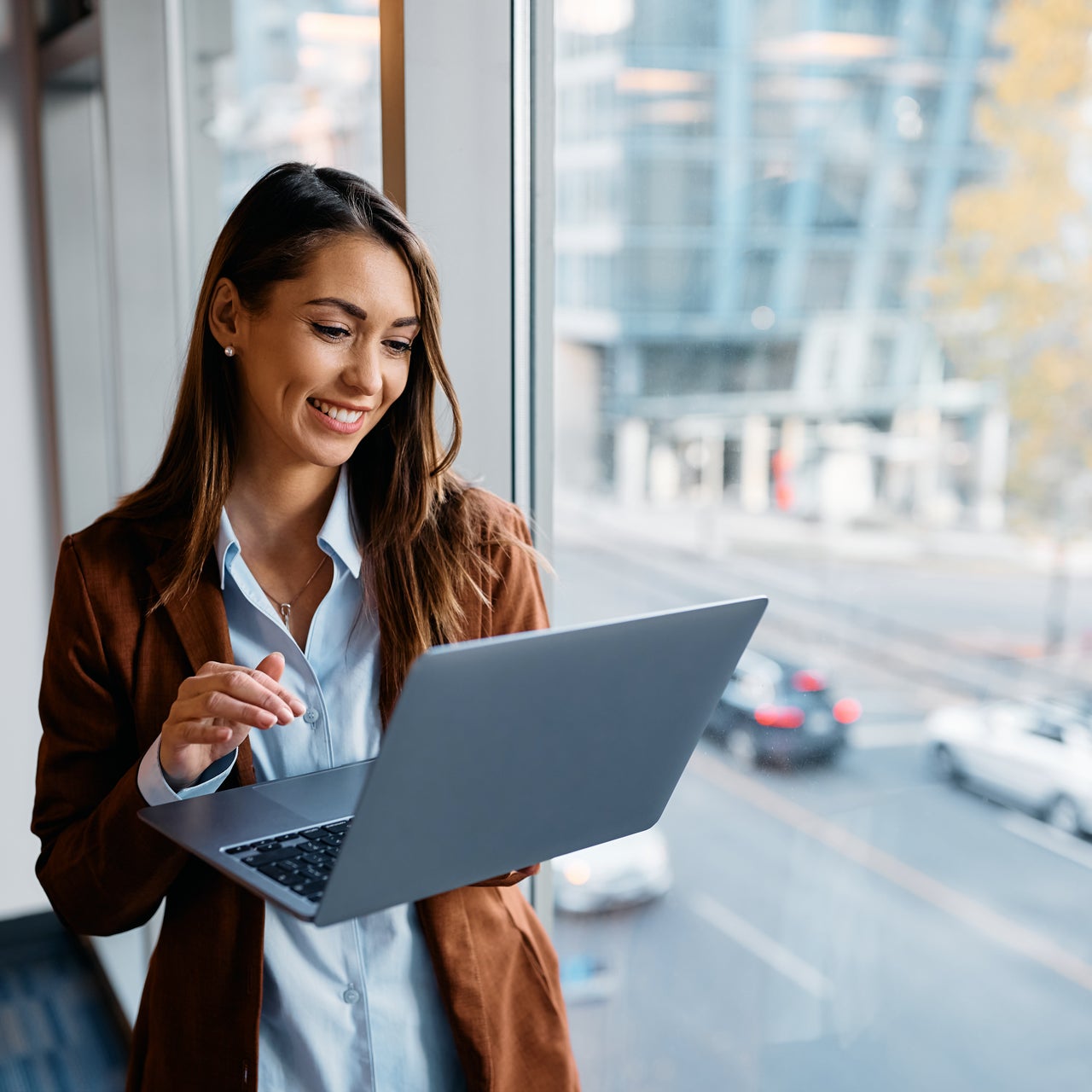 A woman holding a laptop is seen standing near a window.