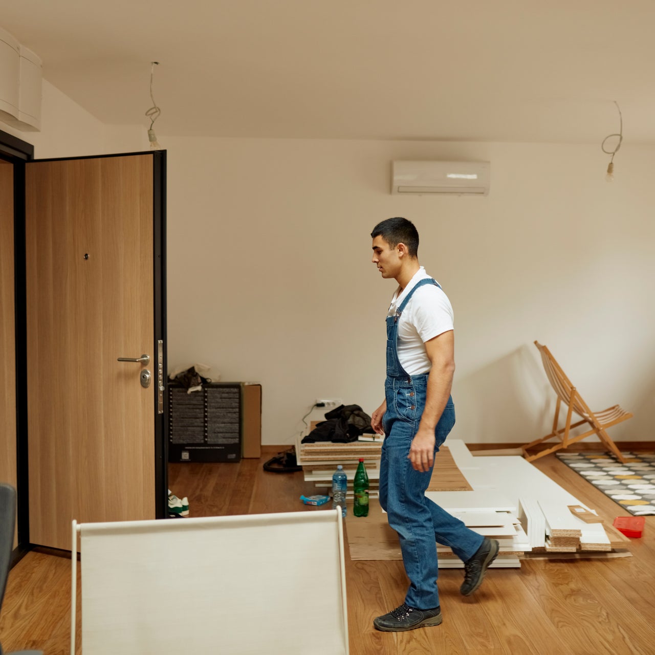 A man in blue overalls and a white t-shirt is walking in a room with furniture parts scattered around.
