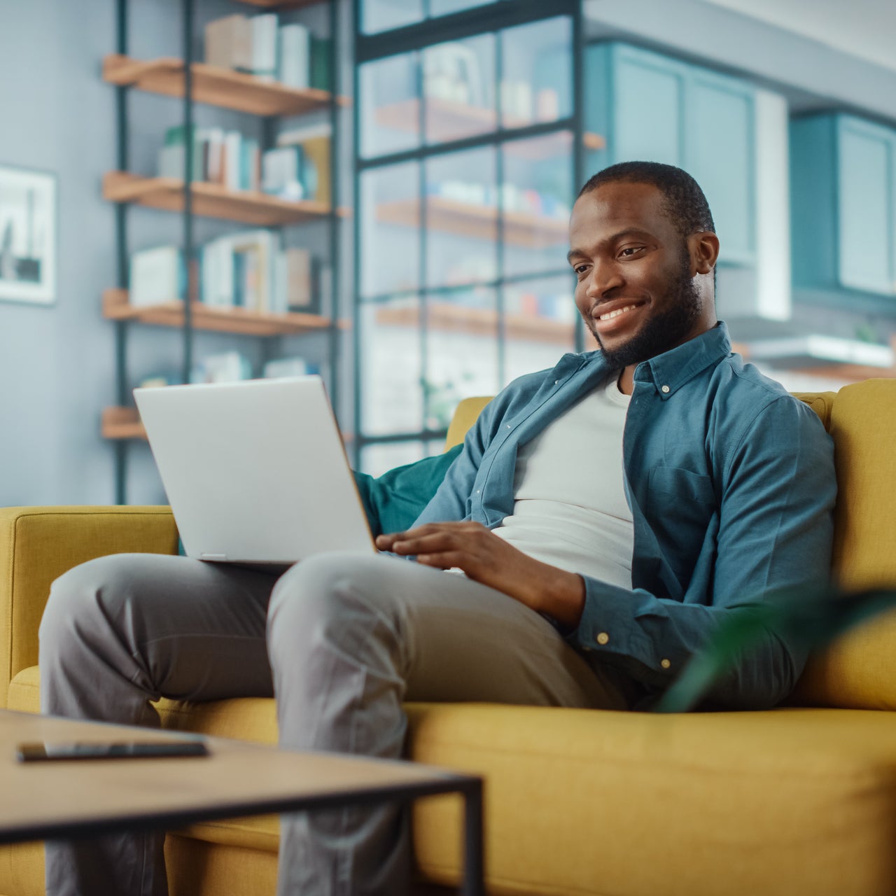 A young man sitting in a relaxed position on his couch and working on his laptop.
