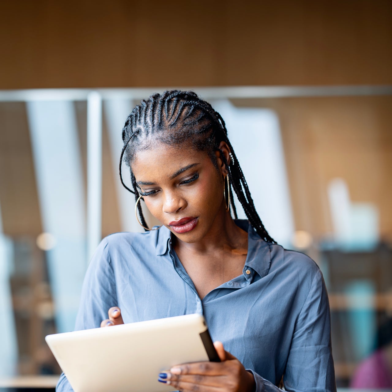 A woman with braided hair, wearing a gray collared shirt and hoop earrings, looking down at a tablet she's holding.