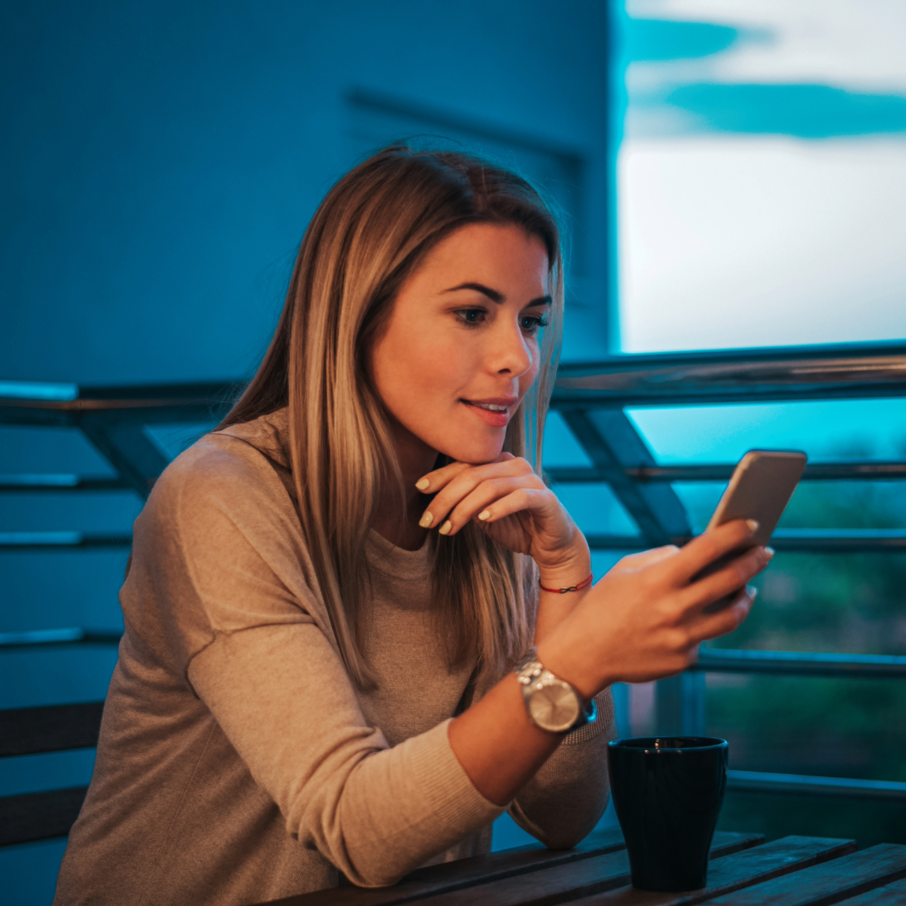 The image shows a woman sitting on a balcony overlooking a city, holding and looking at her phone