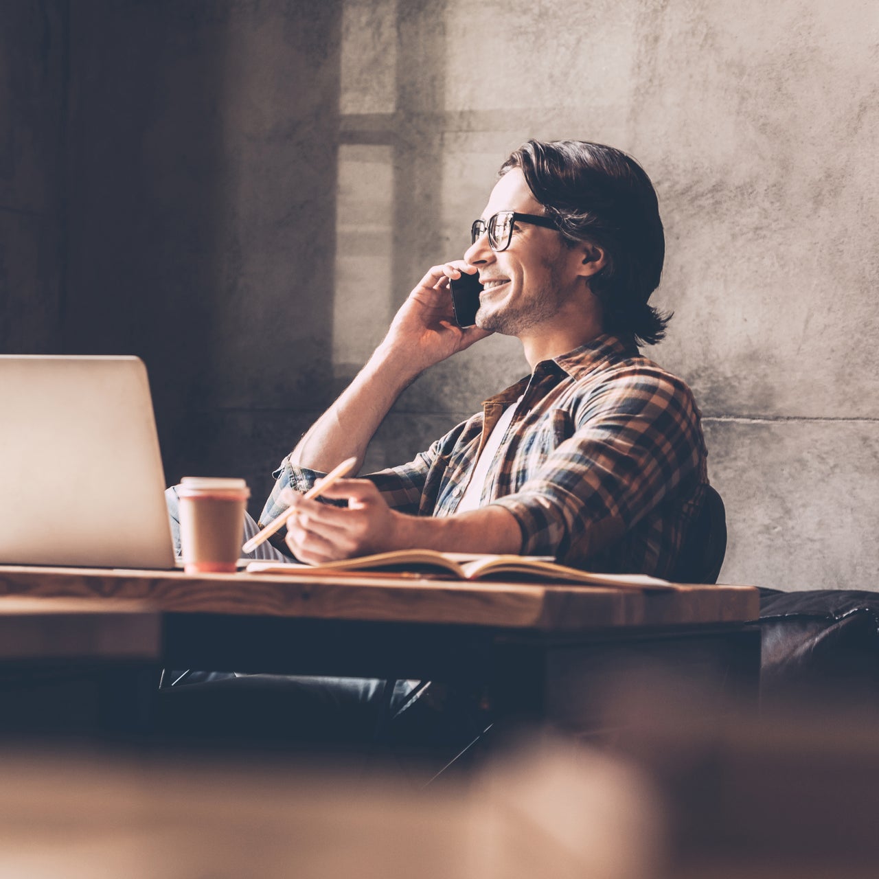 A man sitting on his chair, talking on his mobile phone and smiling. He is on his work desk, as it appears.