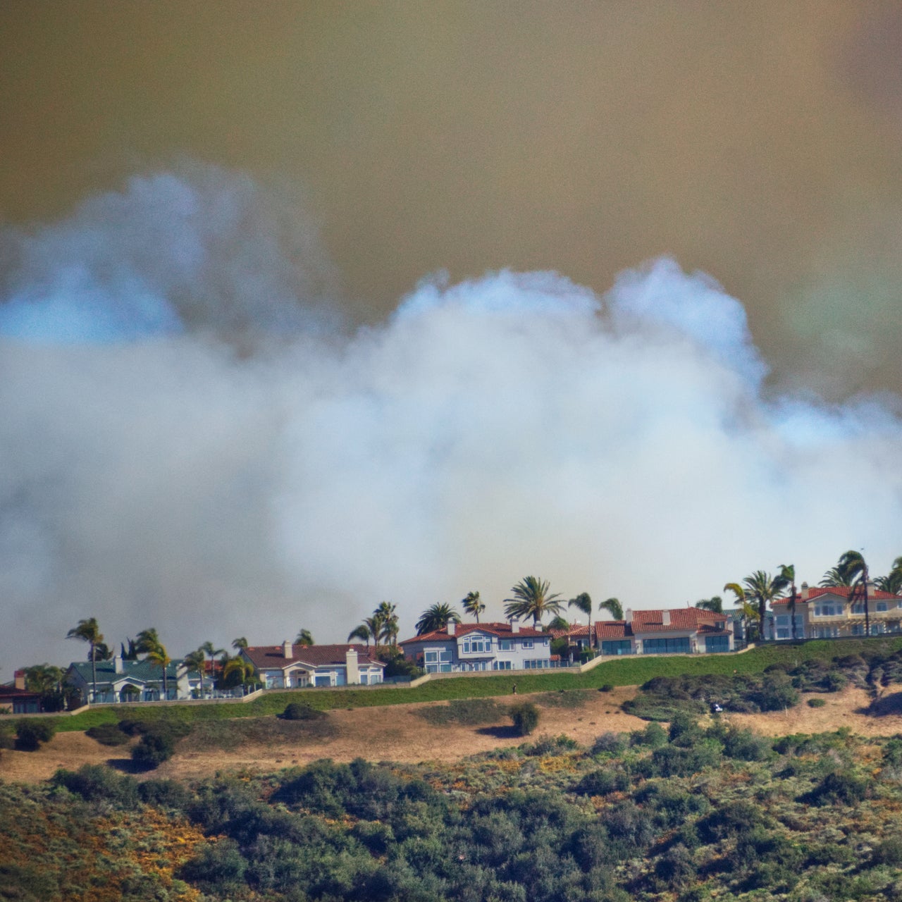 Row of houses on a hill with smoke rising in the background, indicating a possible wildfire hazard.