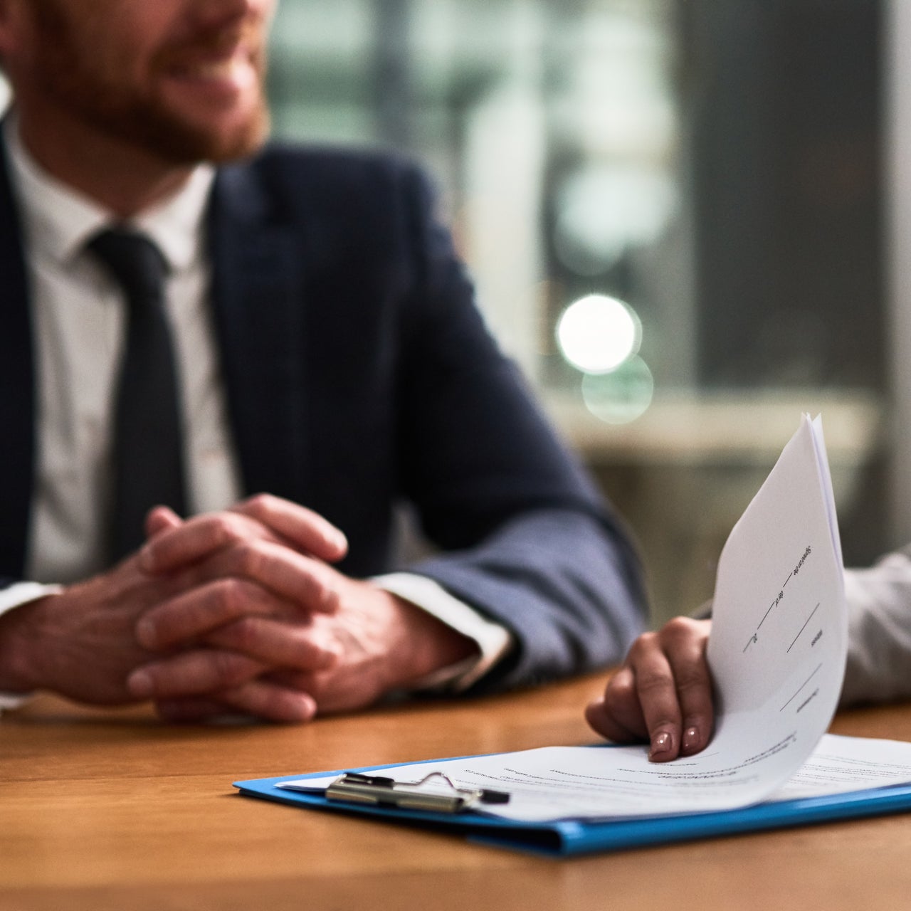 A professional man in a corporate suit sitting on a table with someone whose hand is visible.