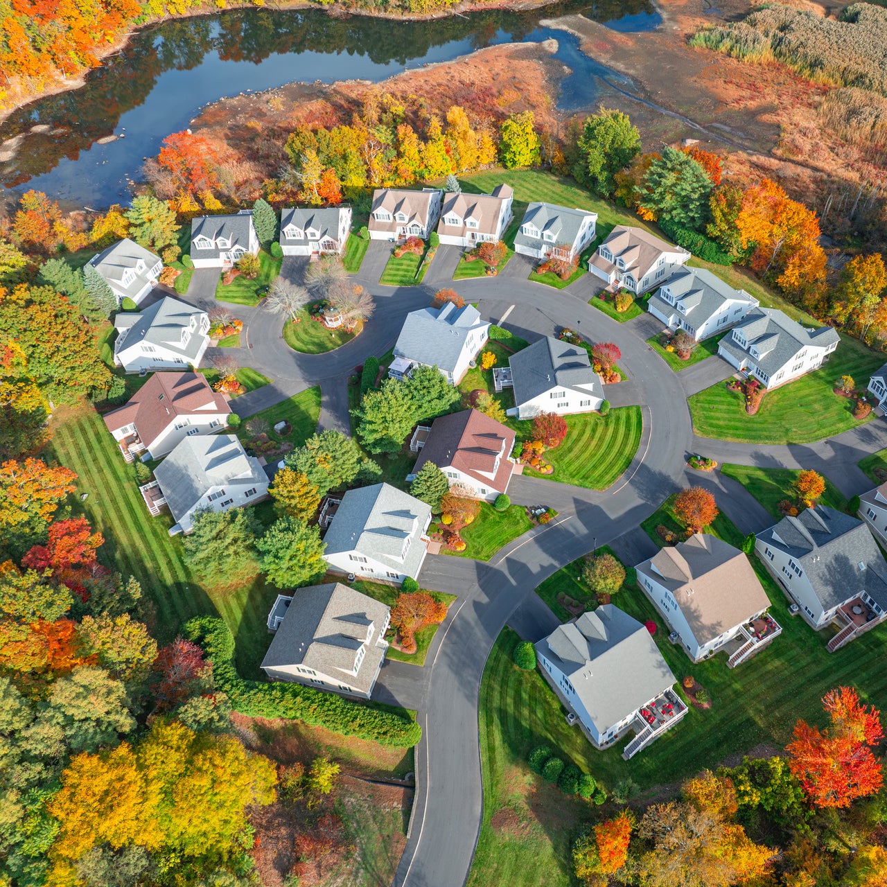 An aerial view of a suburban neighborhood, during the fall season. The houses are arranged in cul-de-sacs and along curved streets, surrounded by trees with vibrant autumn foliage.