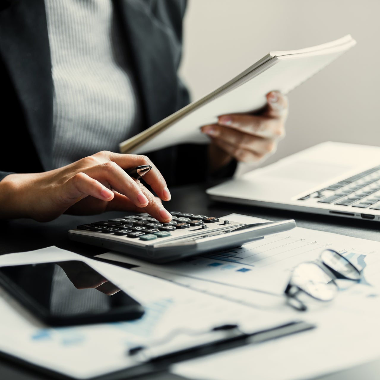 The hands of a professional person is visible. He appears to be in a formal set up, holding a pen and a paper, and working on a calculator kept on desk.