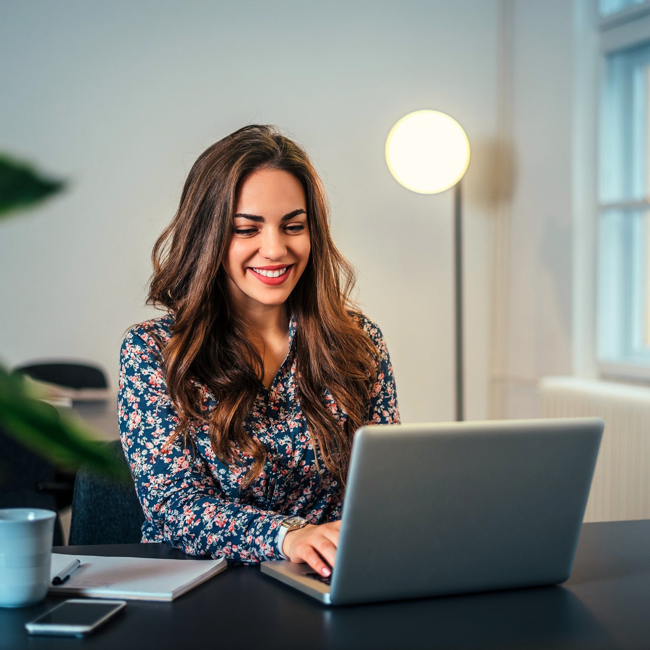 A cheerful professional woman in formal attire is seen working with her laptop kept on her desk.