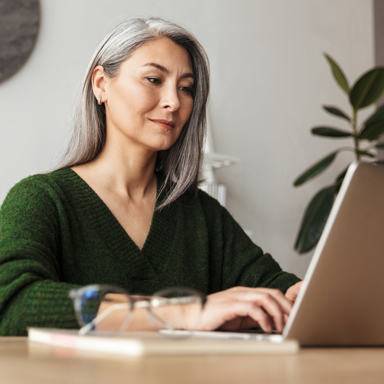 An elderly professional woman in green sweater is working on a laptop kept on a desk.