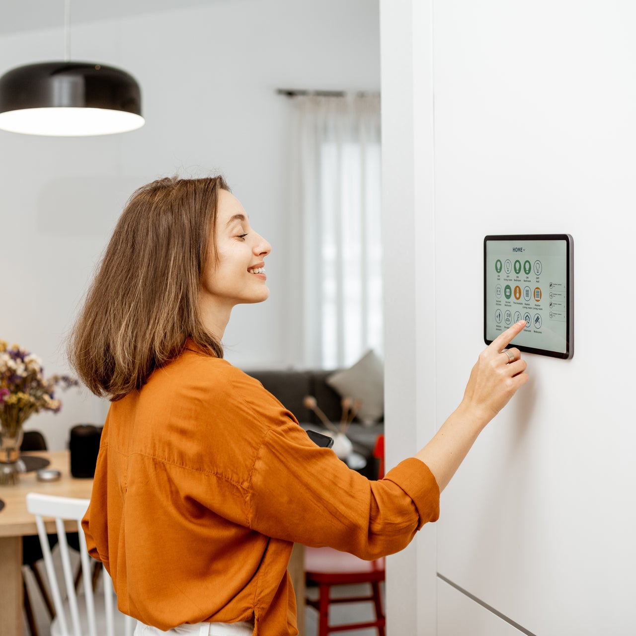 A person interacts with a wall-mounted touchscreen device, possibly using AI, with a house area in the background.